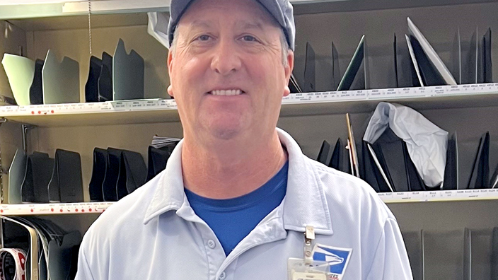 USPS employee wearing blue shorts, a blue shirt and a blue baseball cap standing in front of a sorting station
