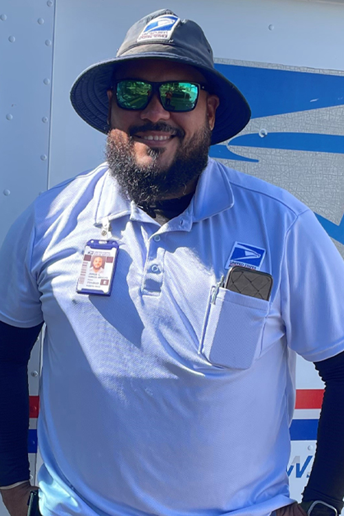 A USPS employee wearing blue shorts, a blue shirt and a blue brimmed hat and standing in front of a Postal Service delivery vehicle