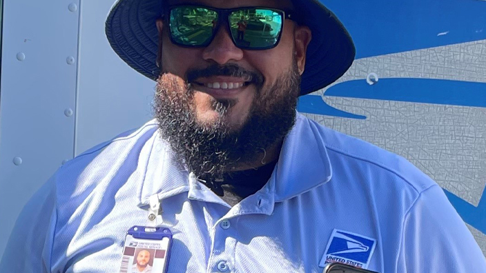 A USPS employee wearing blue shorts, a blue shirt and a blue brimmed hat and standing in front of a Postal Service delivery vehicle