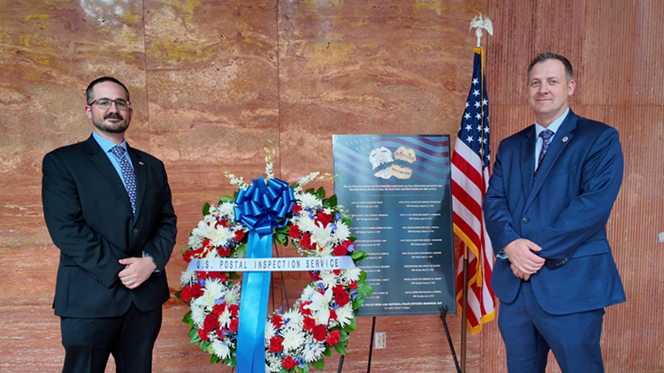 Two postal inspectors standing on either side of a memorial wreath of flowers in USPS headquarters lobby