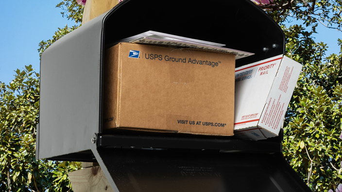 Black mailbox mounted on a wooden post with its door open and displaying a Priority Mail box and a USPS Ground Advantage box
