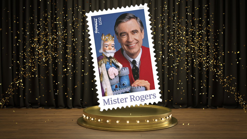 An image of the 2018 USPS Mister Rogers stamp showing a man wearing a red button-up sweater and tie, along with a wooden puppet of a king character.