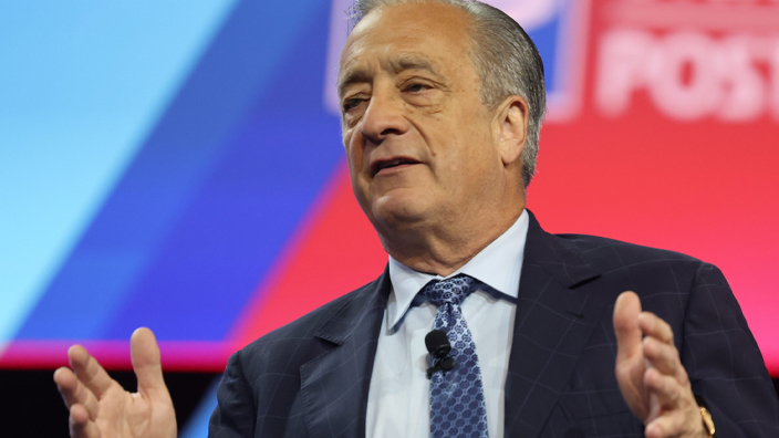 A man in a business suit stands on a stage with the USPS logo looming over his shoulder on a video screen