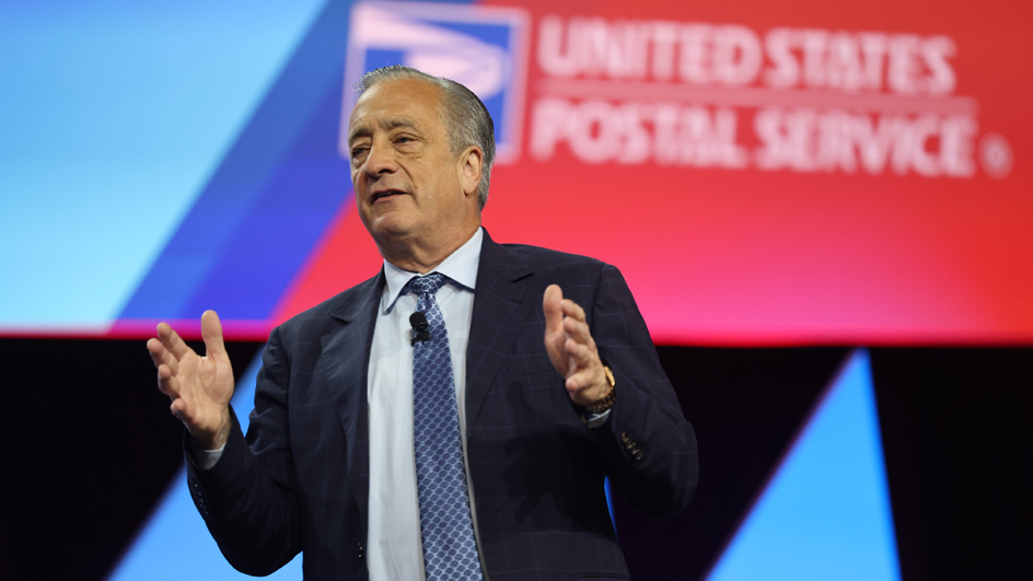 A man in a business suit stands on a stage with the USPS logo looming over his shoulder on a video screen