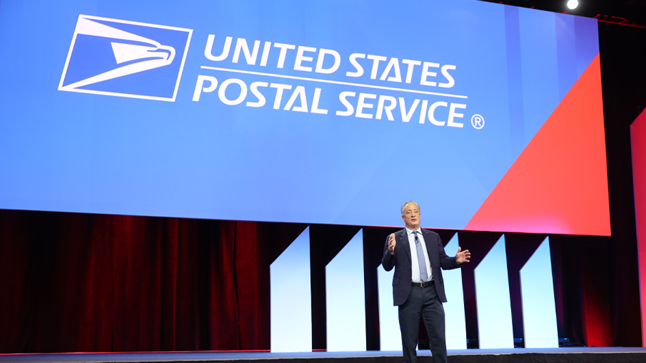A man in a business suit stands on a stage with the USPS logo looming over his head on a video screen