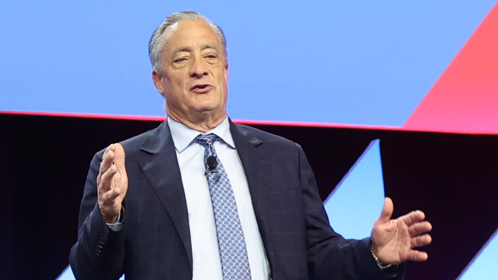 A man in a business suit stands on a stage with the USPS logo looming over his head on a video screen