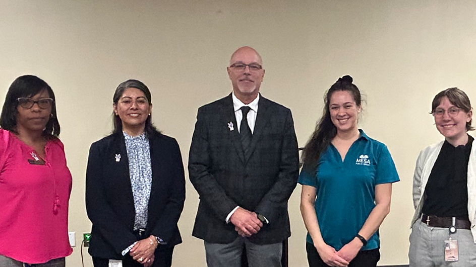 Five people smiling and wearing professional attire standing side by side in a library