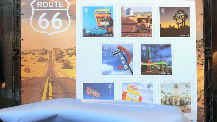 A woman and three men stand on either side of a large image of the Postal Service’s Route 66 stamps