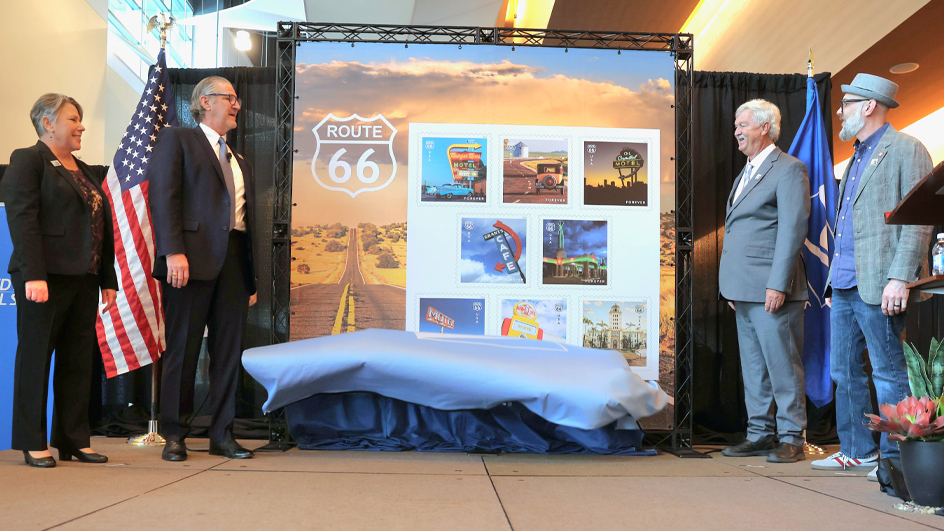 A woman and three men stand on either side of a large image of the Postal Service’s Route 66 stamps
