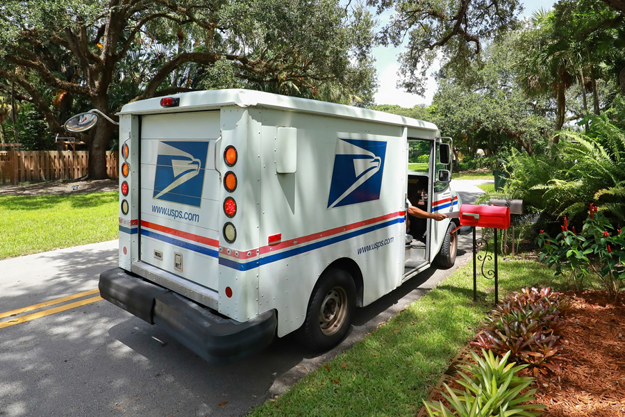 An arm reaches out of USPS delivery vehicle to place letters into a red mailbox