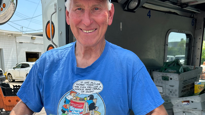 USPS employee in a blue T-shirt and blue hat holding a mail container filled with nonperishable food items
