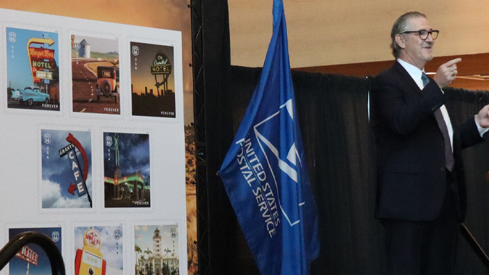 A man in a dark suit gestures from behind a lectern and in front of a large image of the Postal Service’s Route 66 stamps