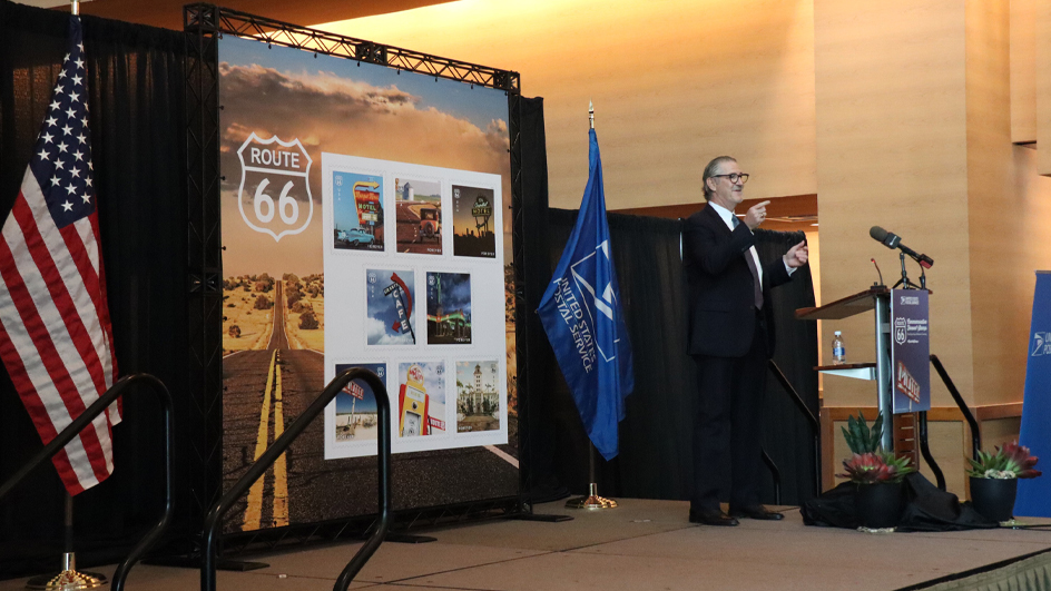 A man in a dark suit gestures from behind a lectern and in front of a large image of the Postal Service’s Route 66 stamps
