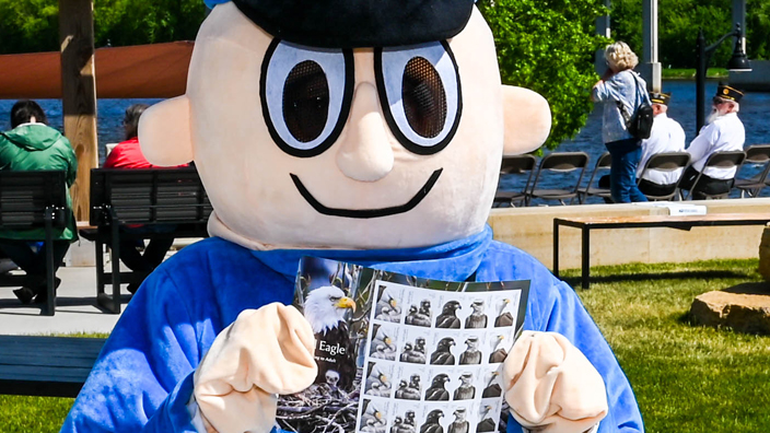 Mr. ZIP mascot wearing a blue letter carrier costume with a mail satchel and holding up a set of Bald Eagle: Hatchling to Adult stamps.