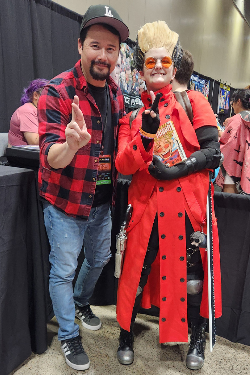 Two people posing at a convention. One wears a red plaid shirt and cap, the other is in a vibrant red costume with goggles.