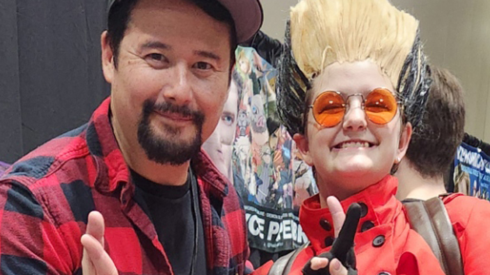 Two people posing at a convention. One wears a red plaid shirt and cap, the other is in a vibrant red costume with goggles.