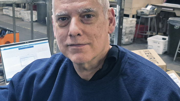 Man seated at a workstation in a mail facility with packages and equipment nearby