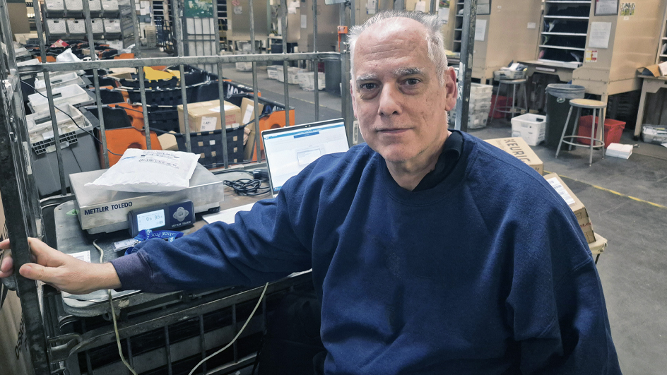 Man seated at a workstation in a mail facility with packages and equipment nearby