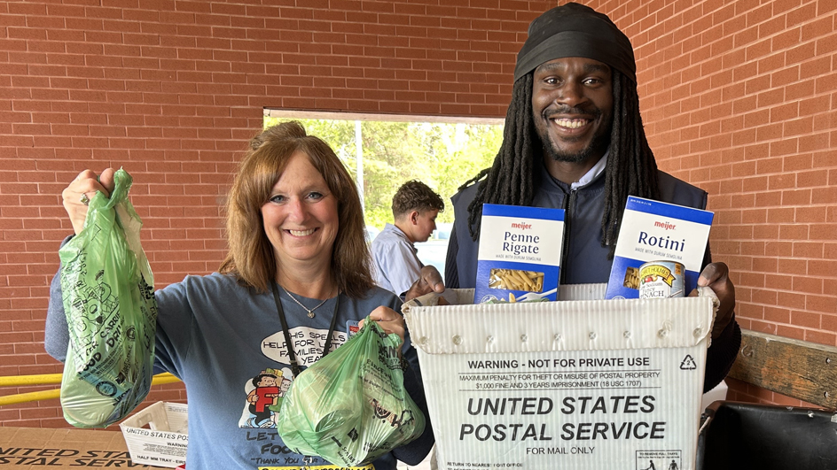 A woman and man hold up bags and a mail container filled with nonperishable food