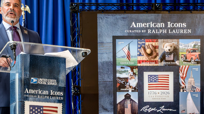 Man in a blue suit standing on stage behind a podium and to the left of an enlarged image of the American Icons stamps pane