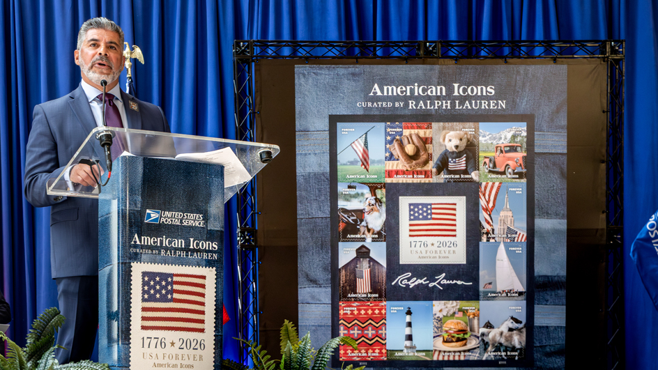 Man in a blue suit standing on stage behind a podium and to the left of an enlarged image of the American Icons stamps pane