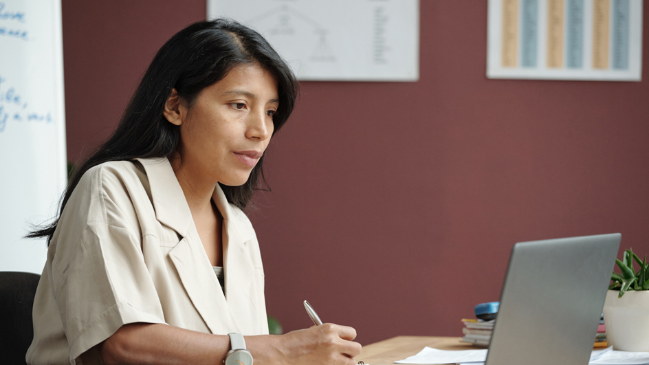 Woman sitting at a desk writing notes while looking a laptop computer screen