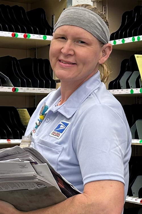 USPS employee in blue shorts and a blue shirt holding a stack of mail