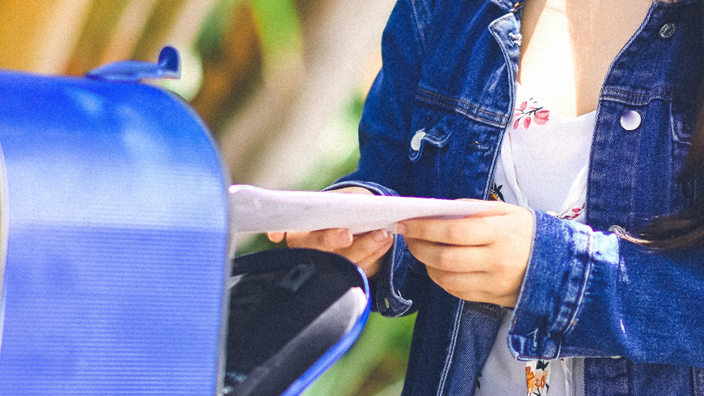 A young woman standing by a blue mailbox and holding a mailpiece