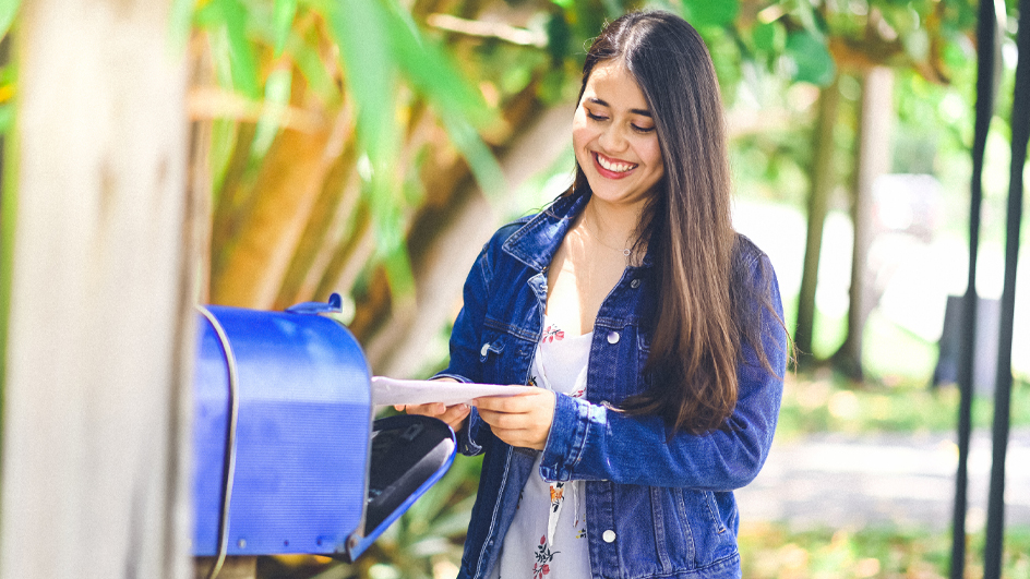 A young woman standing by a blue mailbox and holding a mailpiece