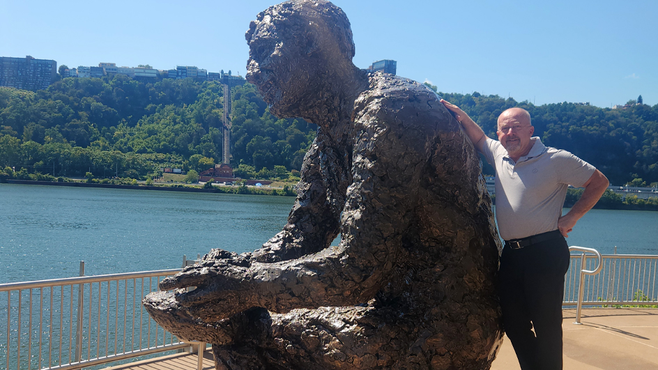 Man standing to the right of a statue of Fred Rogers on a city waterfront
