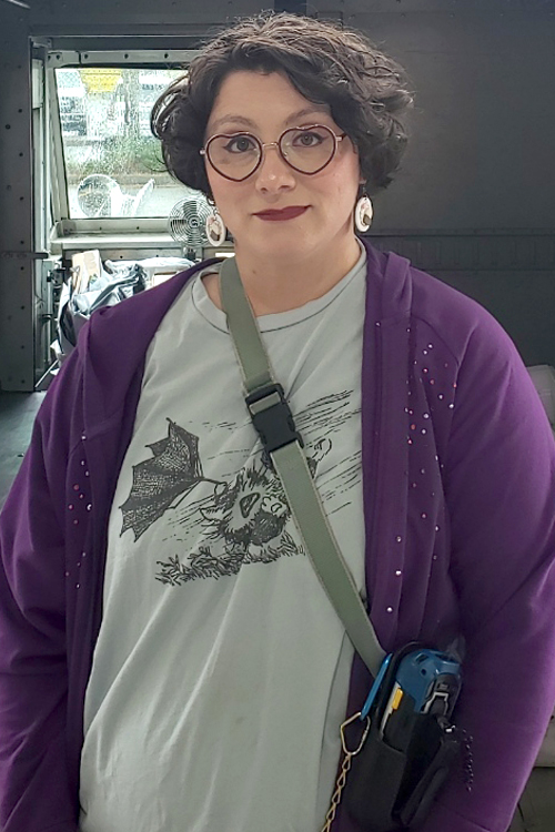 Postal Service employee wearing a purple jacket and standing at the rear of a delivery vehicle