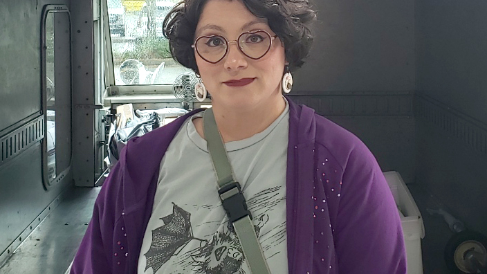 Postal Service employee wearing a purple jacket and standing at the rear of a delivery vehicle