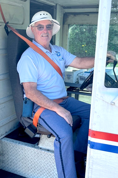 SPS employee wearing a blue uniform and light-colored hat and sitting in driver’s seat of a delivery vehicle with seat belt buckled