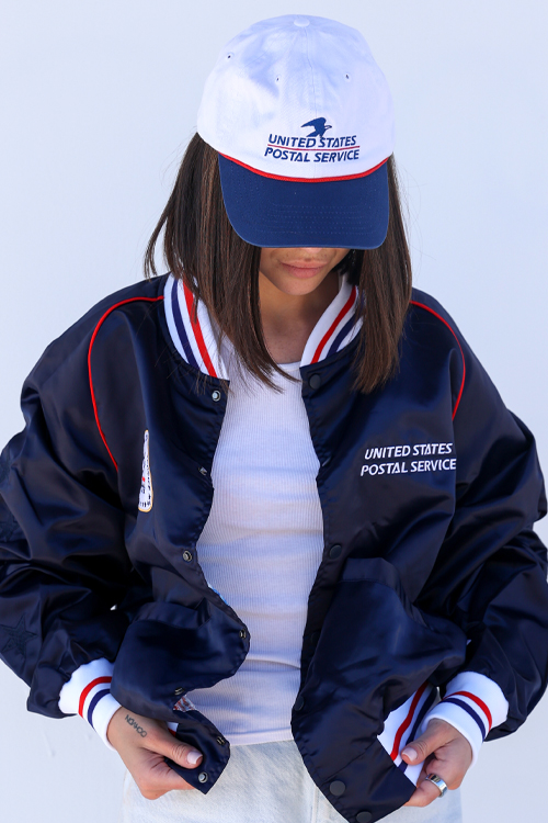 A person standing against a white backdrop and modeling a white USPS-branded baseball hat and a blue USPS-branded jacket