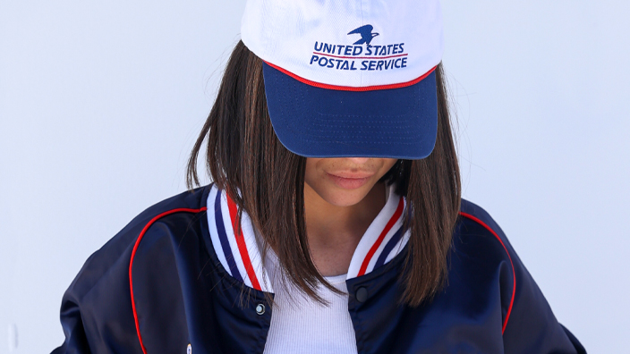 A person standing against a white backdrop and modeling a white USPS-branded baseball hat and a blue USPS-branded jacket