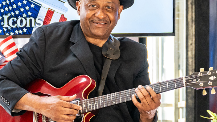 A man dressed in black and perched on a stool holds a red guitar at the American Icons stamp ceremony