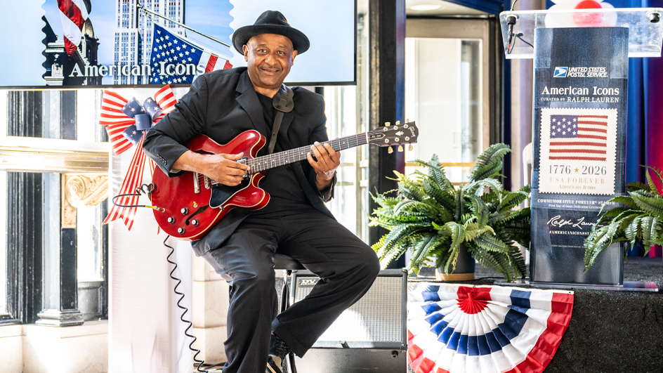 A man dressed in black and perched on a stool holds a red guitar at the American Icons stamp ceremony