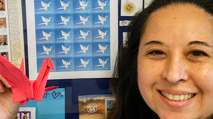USPS employee holding up a red origami crane next to a pane of the International Peace stamps