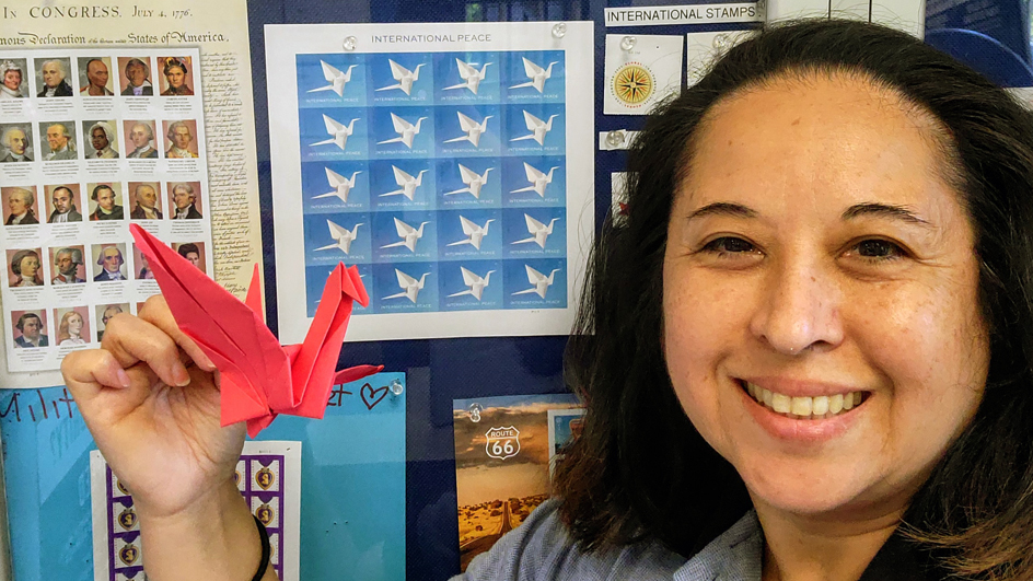 USPS employee holding up a red origami crane next to a pane of the International Peace stamps