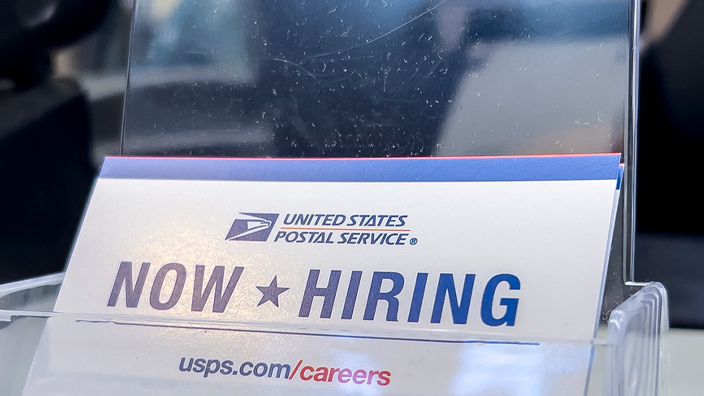 USPS counter sign displaying a “Now Hiring” card