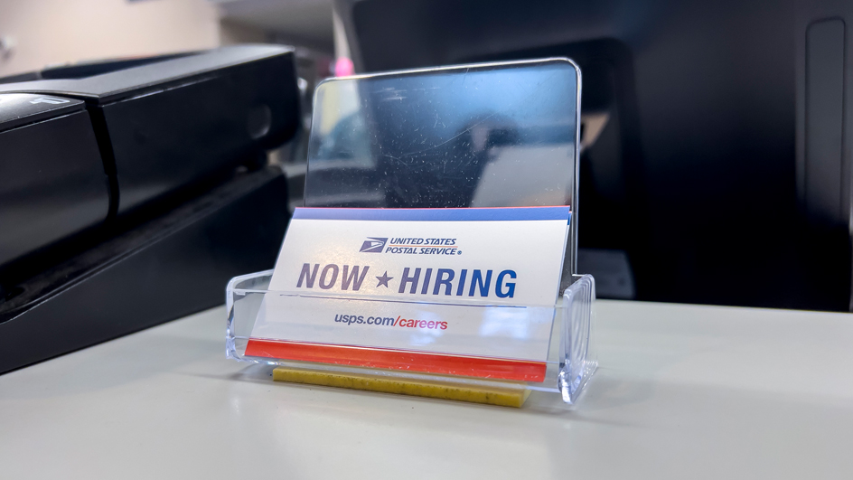 USPS counter sign displaying a “Now Hiring” card