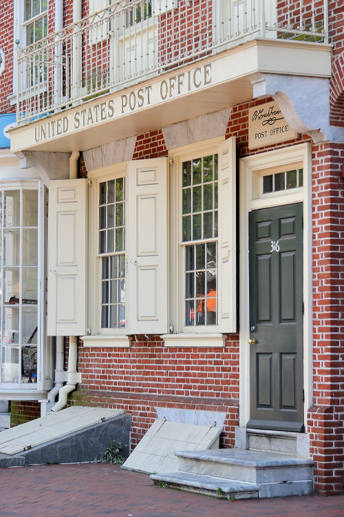 The front of a red brick building with a balcony displaying a sign saying “United States Post Office”