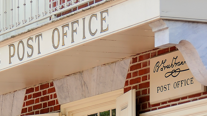 The front of a red brick building with a balcony displaying a sign saying “United States Post Office”
