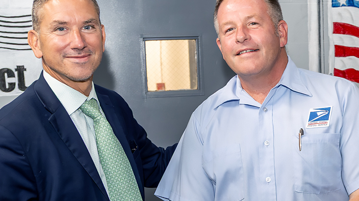 A man in a blue suite and light green tie stands with another man wearing a light blue short-sleeve shirt with a USPS patch.