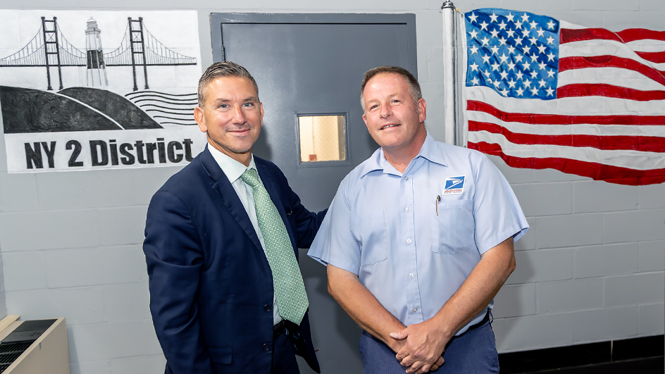 A man in a blue suite and light green tie stands with another man wearing a light blue short-sleeve shirt with a USPS patch.