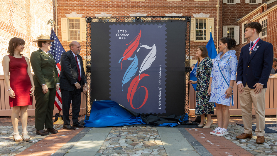 Three ceremony participants stand on each side of an enlarged image of the Declaration of Independence stamp