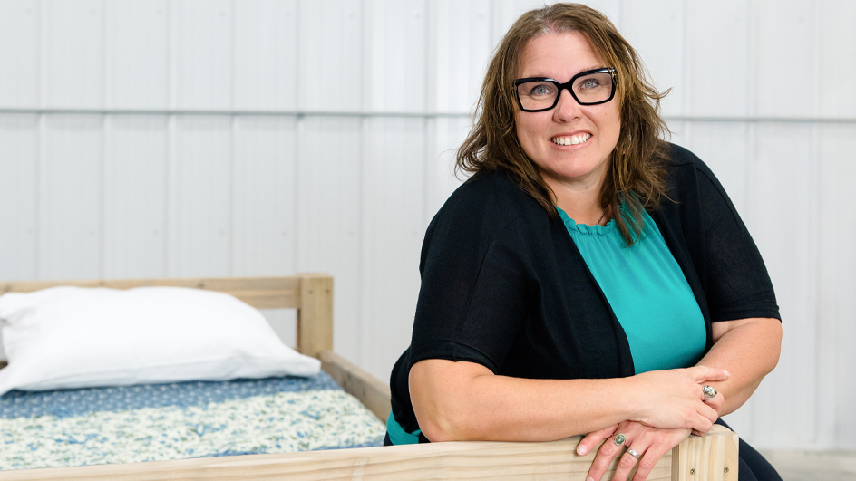 A woman in a black sweater and turquoise blouse sits on a wood-frame bed