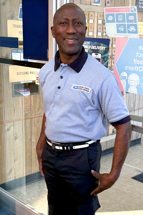 USPS employee wearing black pants and a light-colored polo shirt and standing in front of a glass door in a Post Office lobby