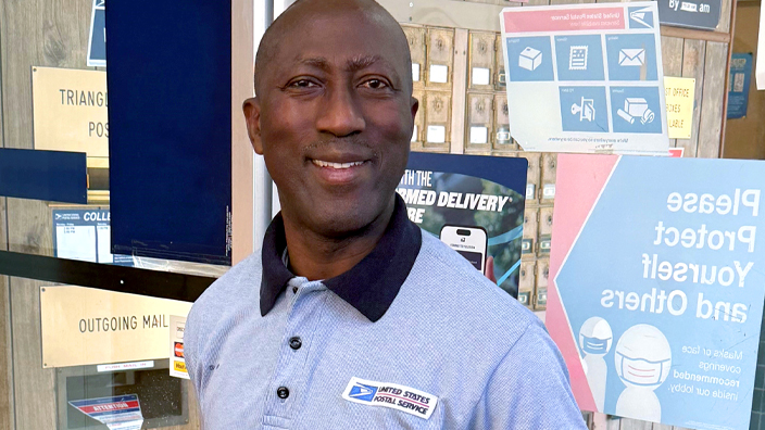 USPS employee wearing black pants and a light-colored polo shirt and standing in front of a glass door in a Post Office lobby