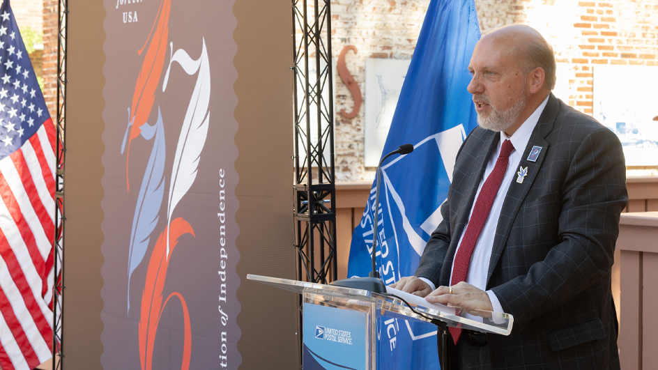 A man in a dark suit speaks from behind a lectern with a large image of the Declaration of Independence stamp off to his side.